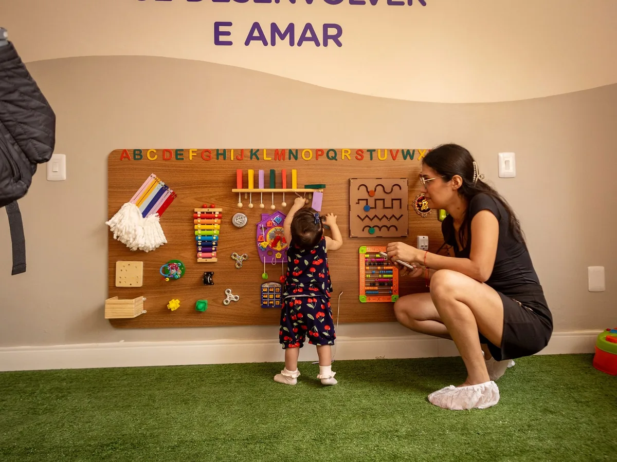 Criança pequena explora um painel sensorial de madeira com letras, xilofone e peças de encaixe, acompanhada pela mãe