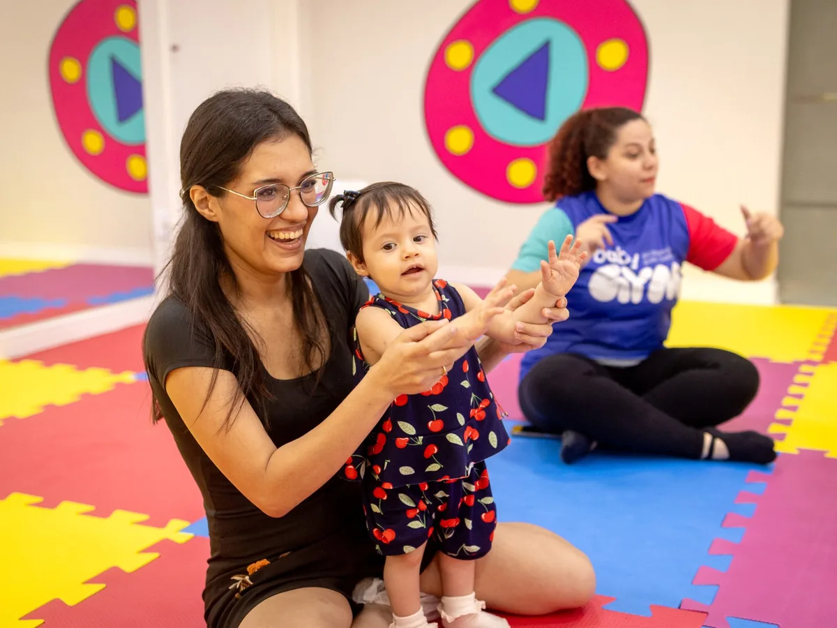 Mãe sentada no tatame segura a filha pequena pelas mãos durante uma atividade, com uma professora da Baby Gym ao fundo